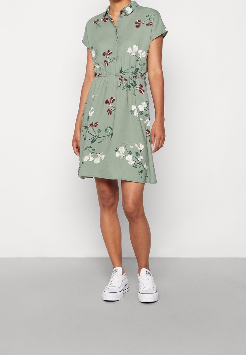 Robe florale verte avec un col, des manches courtes et une taille élastique. Motifs de fleurs blanches et accents bordeaux. Portée avec des baskets blanches.