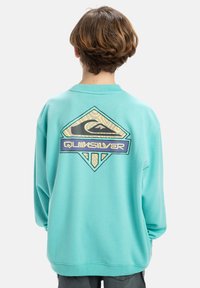 Törökzöld pulóver, amelyen egy nagy, gyémánt alakú Quiksilver logó található hullám grafikával és törzsi mintákkal a hátsó részen. Puha anyag, oversized fazon.