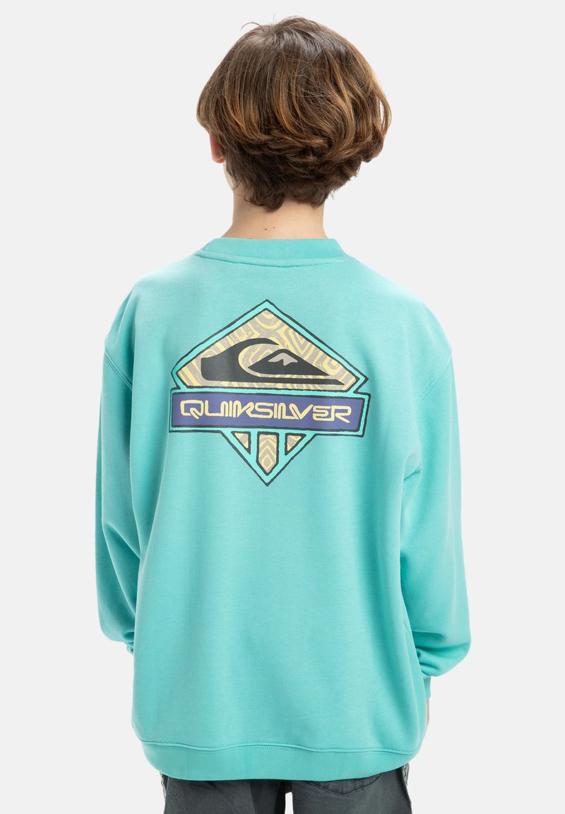 Törökzöld pulóver, amelyen egy nagy, gyémánt alakú Quiksilver logó található hullám grafikával és törzsi mintákkal a hátsó részen. Puha anyag, oversized fazon.