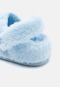 Világoskék, bolyhos papucs, puha textúrával, keresztpántokkal és lekerekített dizájnnal. A talpbetét párnázott a kényelem érdekében.