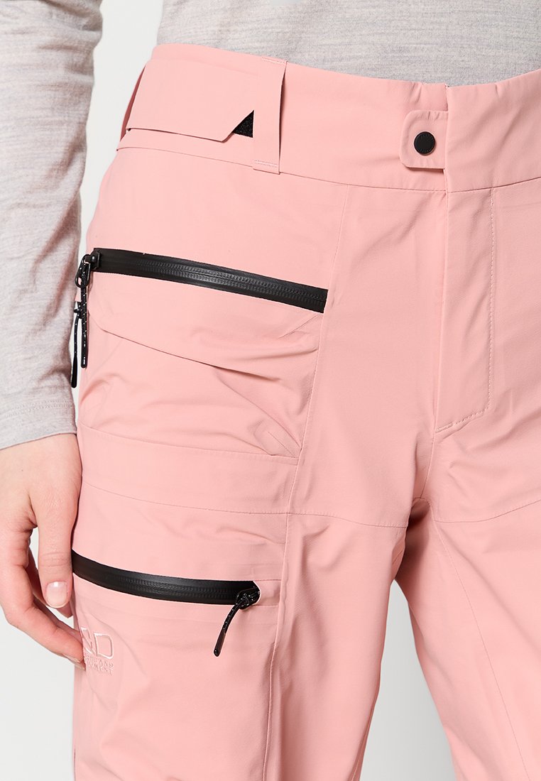 Nahaufnahme einer Person, die pinke Hosen mit zwei schwarzen Reißverschlusstaschen trägt und die Seite der Hose in der Nähe der unteren Tasche hält.