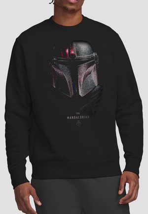 Mann trägt einen schwarzen Sweatshirt mit einer detaillierten silbernen Mandalorian-Helm-Grafik und dem Schriftzug „The Mandalorian“ darunter.