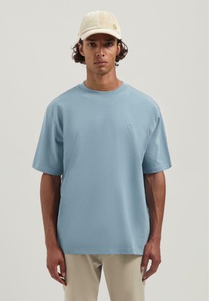 Junger Mann in einem hellblauen übergroßen T-Shirt, beigen Hosen und einer cremefarbenen Cord-Baseballmütze, der vor einem schlichten Hintergrund steht.