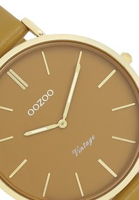 Montre ronde marron avec des accents dorés, aiguilles des heures et des minutes dorées, et marqueurs dorés. Équipée d'un bracelet en cuir marron. Le texte indique "oozoo Vintage."