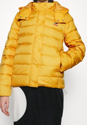 Personne portant une doudoune matelassée jaune avec un devant à boutons-pression et un petit logo tricolore sur la poitrine.