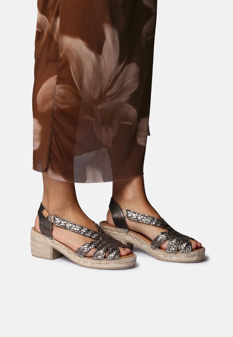 Jalad, mis kannavad metallik-hõbedase punutud rihmaga platvorm-sandaleid koos kootud espadrille-baseeruvate talladega, sobitatud pruunide lillemustrilise läbipaistva kangaga seelikuga.