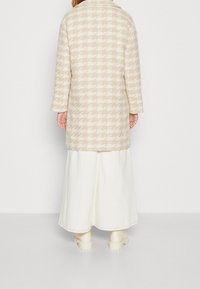 Beige kappa med hundtandsmönster, långärmad, med krage och midilängd, bärs över en vit kjol och ljusa skor.