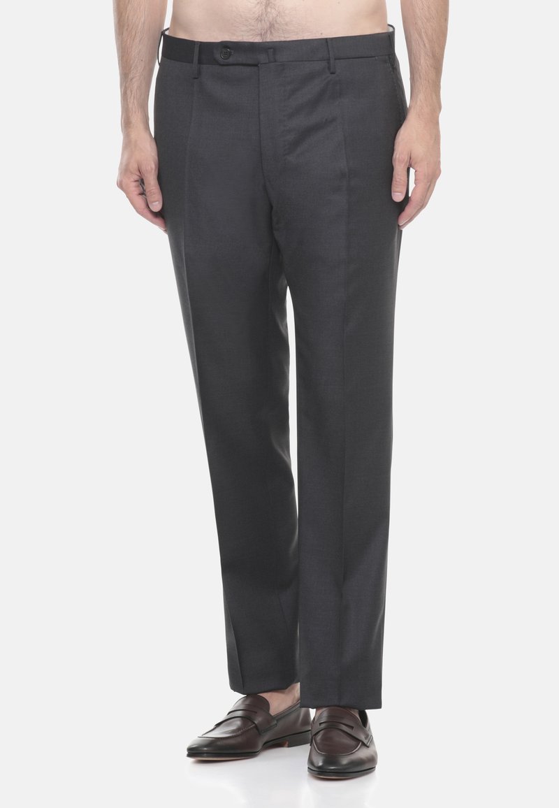 Uomo che indossa pantaloni formali grigio scuro e mocassini in pelle marrone scuro, in piedi davanti a uno sfondo bianco semplice.