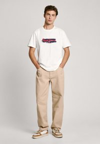 Camiseta blanca de algodón con un estampado gráfico de colores, combinada con pantalones beige y zapatillas con detalles marrones. Corte simple y relajado.