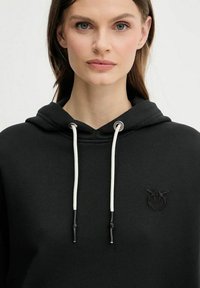 Donna che indossa un felpa nera con cappuccio, con cordini bianchi e un logo ricamato nero sul lato sinistro del petto, che guarda dritto davanti a sé su uno sfondo chiaro.