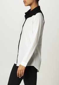 Blouse blanche à manches longues avec un col en dentelle noire, fermeture à boutons sur le devant et bordure noire contrastante. Texture lisse et coupe régulière.