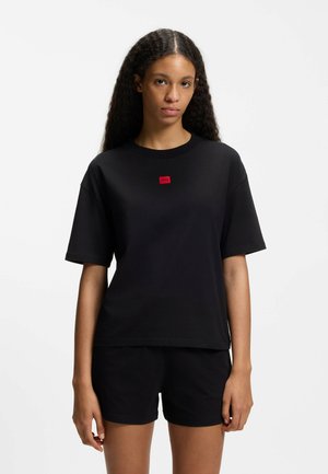 Jeune femme aux longs cheveux bouclés porte un T-shirt noir uni oversize avec un petit logo rouge et un short noir, debout devant un fond blanc.