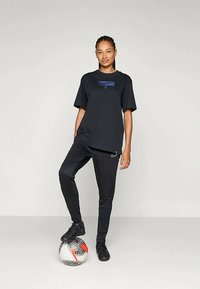 Černé sportovní tričko s modrým logem, v kombinaci s černými kalhotami. Model stojí na vzorovaném fotbalovém míči a má na sobě černé tenisky.