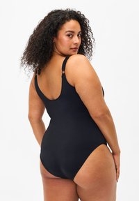 Vrouw met krullend haar die een zwart eendelig badpak draagt, kijkt over haar schouder tegen een effen witte achtergrond.