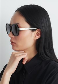 Ochelari de soare de mari dimensiuni, cu un cadru lucios, având brațe accentuate cu argint, expuși pe un model cu păr drept de culoare închisă.