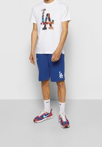 T-shirt en coton blanc avec un logo coloré de Los Angeles, accompagné d'un short de sport bleu avec une impression blanche LA, et des baskets rouges et bleues.