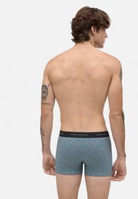 Hombre con tatuajes que lleva puesta una boxer azul con un patrón y cinturilla negra, de pie de espaldas contra un fondo blanco.