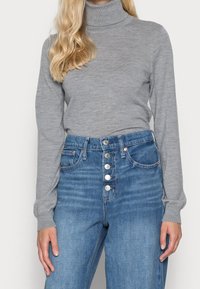 Pull à col roulé gris avec texture côtelée, associé à un jean denim bleu taille haute avec fermeture à cinq boutons et poches avant.
