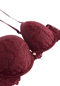 rega di pizzo bordeaux con coppe imbottite, sovrapposizione di pizzo floreale, spalline regolabili e un piccolo accento di rosa rosa sulla fascia. Materiali texturizzati e lisci.