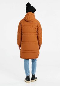 Orange pufferjakke med hætte, med horisontale hætte og en mellemlang design. Bæret med lyseblå jeans og sorte ankelstøvler.