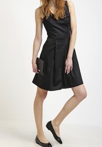 Musta hihaton mekko, jossa on vartalonmyötäinen yläosa ja levenevä helma. Malli pitää mustaa clutch-laukkua ja käyttää mustia tasapohjaisia kenkiä. Kangas on sileää.