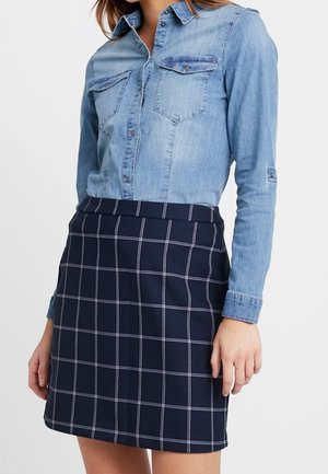 Kobieta nosząca jasno-niebieską koszulę dżinsową włożoną do granatowej spódnicy w biały wzór kratki typu „windowpane”.
