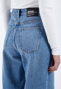 Light blue denim jeans with a high waist, five pockets, and subtle fading. Features a black brand label on the back waistband.