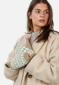 Frau in einem beigefarbenen Wollmantel, die beigen, weißen und mintfarbenen gemusterten Handschuhe trägt und zur Seite blickt, vor einem schlichten Hintergrund.