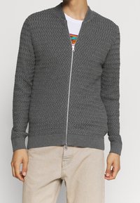 Grå zip-up kofta med texturerad stickad mönster, ribbade ärmslut och nederkant. Har en silverfärgad dragkedja och stående krage, bärs över en vit skjorta.