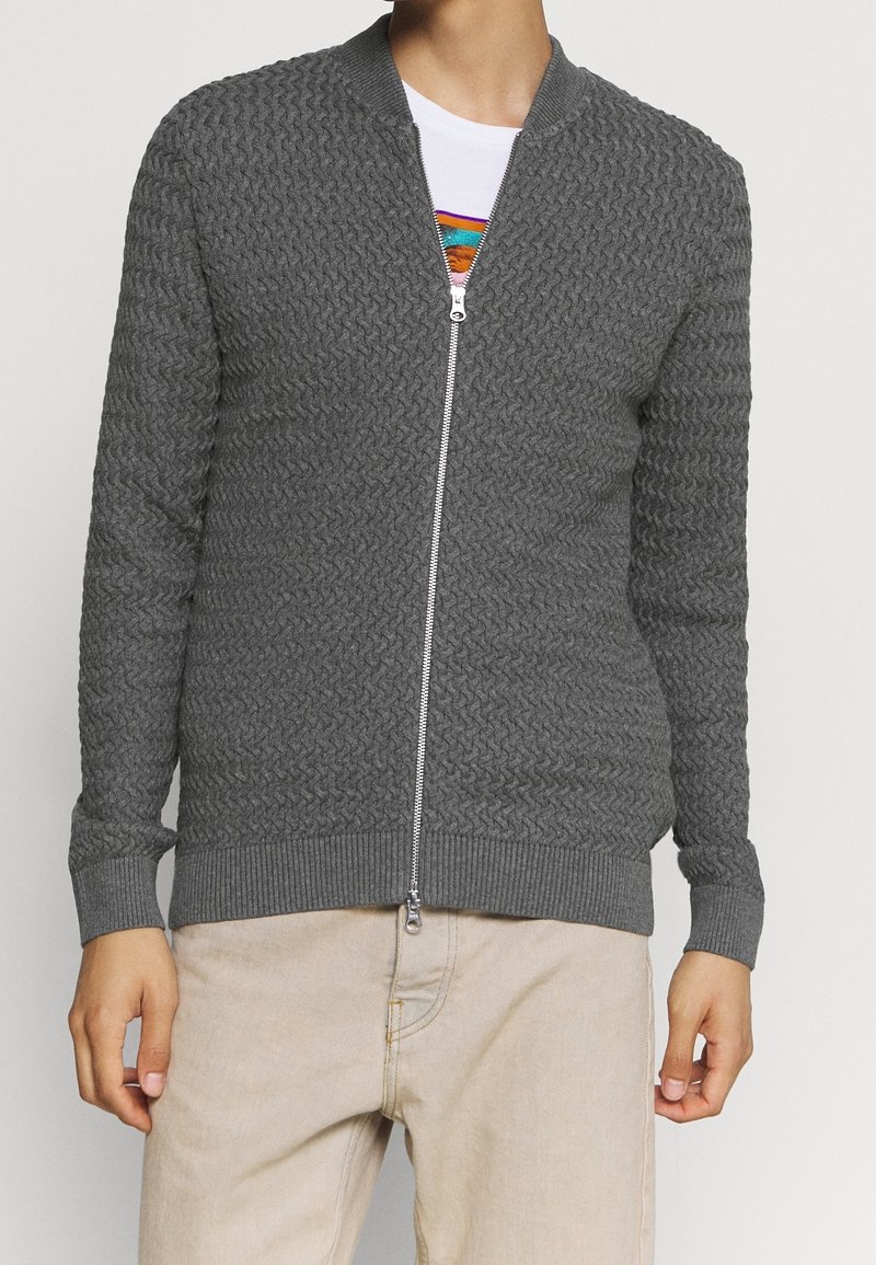 Grå zip-up kofta med texturerad stickad mönster, ribbade ärmslut och nederkant. Har en silverfärgad dragkedja och stående krage, bärs över en vit skjorta.