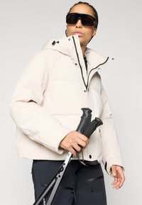 Μπεζ μόνωτη τζάκετ με ψηλό γιακά, μαύρο φερμουάρ και κουκούλα με κορδόνια. Το μοντέλο κρατάει μπαστούνια πεζοπορίας, φορώντας μεγάλα μαύρα γυαλιά ηλίου.