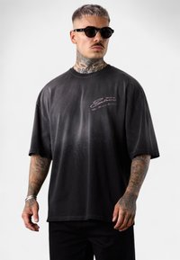 Black gradient short-sleeve t-shirt with a round neck and pink embroidered logo on the left chest. Model wears black sunglasses and patterned tattoos.