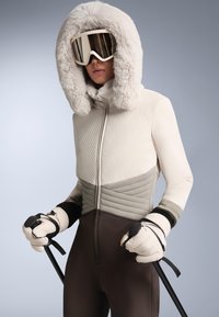 Fehér és szürke szigetelt sídzseki műszőrmés kapucnival, párnázott kesztyűkkel, sötét sí nadrággal és síbotokkal. Simább textúrájú, modern kialakítás.