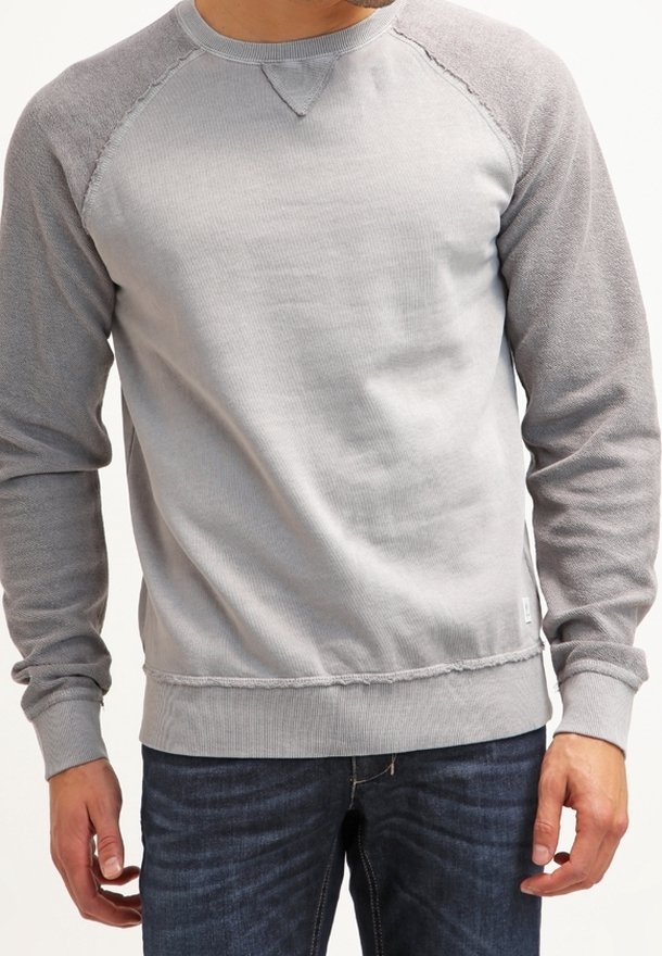 Homme portant un sweat-shirt gris clair à col rond avec des manches raglan gris plus foncé et un jean bleu foncé, debout avec les bras détendus.