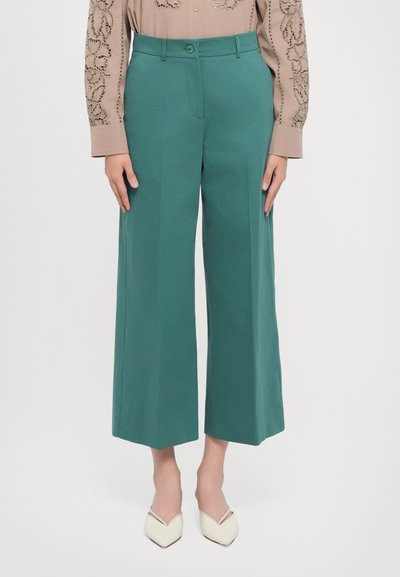 Türkisfarbene Weitleg-Hosen mit tailliertem Schnitt, glattem Vorderteil und weichem Material. Kombiniert mit einer beigen gemusterten Bluse und cremefarbenen spitzen Schuhen.