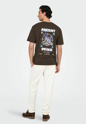 Bruine katoenen T-shirt met een kompass ontwerp en bergafbeeldingen in blauw en wit, tekst "ASCENT PEAKS," gecombineerd met crèmekleurige broeken.