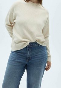 Suéter de punto beige con cuello redondo, combinado con pantalones vaqueros azul de tiro alto. El suéter tiene un ajuste ligeramente holgado y un dobladillo acanalado.