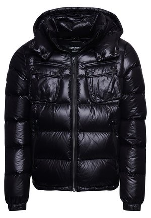 Personne portant une doudoune noire brillante, un pantalon de survêtement noir avec un petit logo, des baskets blanches, debout devant un mur et un sol blancs.
