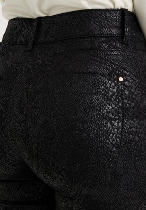 Pantalon noir texturé avec un motif de peau de reptile. Il comporte une taille ajustée et un petit accent doré près de la poche arrière. Matière douce et brillante.
