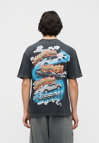 Άνδρας που φοράει μαύρο T-shirt με πολύχρωμες απεικονίσεις του αυτοκινήτου Nissan Skyline GT-R και κείμενο στην πλάτη, στέκεται μπροστά σε απλό φόντο.