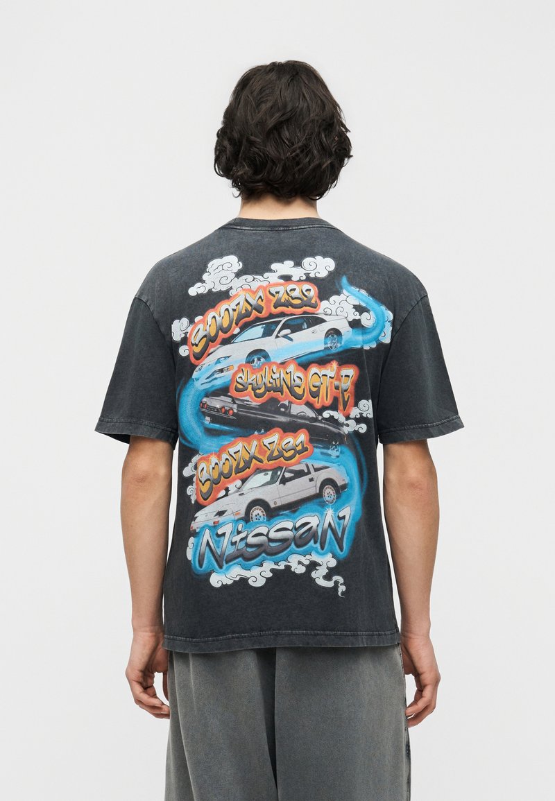 Άνδρας που φοράει μαύρο T-shirt με πολύχρωμες απεικονίσεις του αυτοκινήτου Nissan Skyline GT-R και κείμενο στην πλάτη, στέκεται μπροστά σε απλό φόντο.