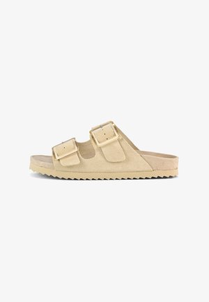 Beige suede slide sandal featuring two adjustable straps with rectangular buckles and a textured, serrated sole.