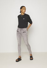T-shirt preto de manga curta com logótipo branco, combinado com jeans cinzentos de corte ajustado com bolsos com fechos éclair, e sandálias pretas rasas.