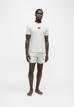 Homme adulte debout pieds nus portant une chemise à manches courtes gris clair et un short assorti avec un logo rouge Hugo sur la poitrine.