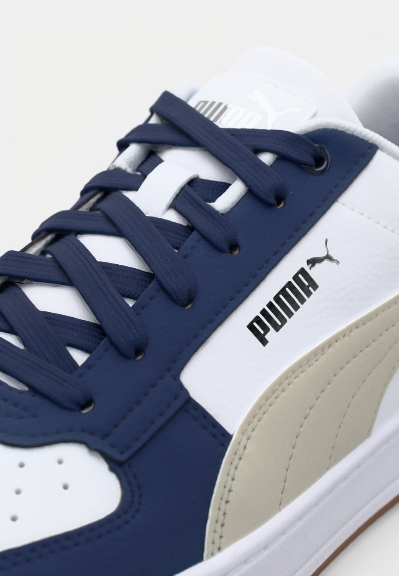 Baskets sportives avec une tige en cuir bleu marine et blanc, accentuée par du beige. Marquage noir sur le côté, lacets plats et bout perforé.