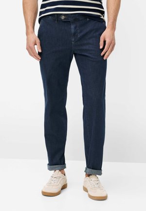 Mand iført mørkeblå op-rullede jeans, hvide sneakers med gummisåler og en marineblå og hvidstribet skjorte, stående mod en hvid baggrund.