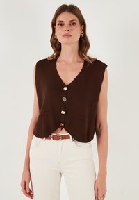 Brązowy sweter bez rękawów z dekoltem w kształcie V, ozdobiony trzema złotymi guzikami i dwiema przednimi kieszeniami, zestawiony z białymi spodniami i brązowym paskiem.