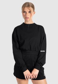 Zwarte sweater met lange mouwen en een ronde halslijn, met een gather taille en wit logo detail. Gekleed met zwarte shorts.