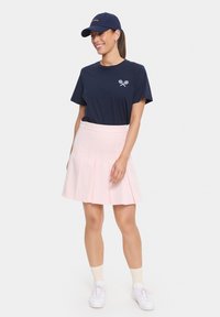 Navy t-shirt with crossed tennis racquets logo, pink pleated skirt, white sneakers, and navy cap with embroidered text. Smooth fabric, casual style.