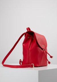 Mochila de imitación cuero rojo con forma redondeada, cierre con solapa, correa para el hombro ajustable y acentos de hardware en tono dorado. Textura suave.